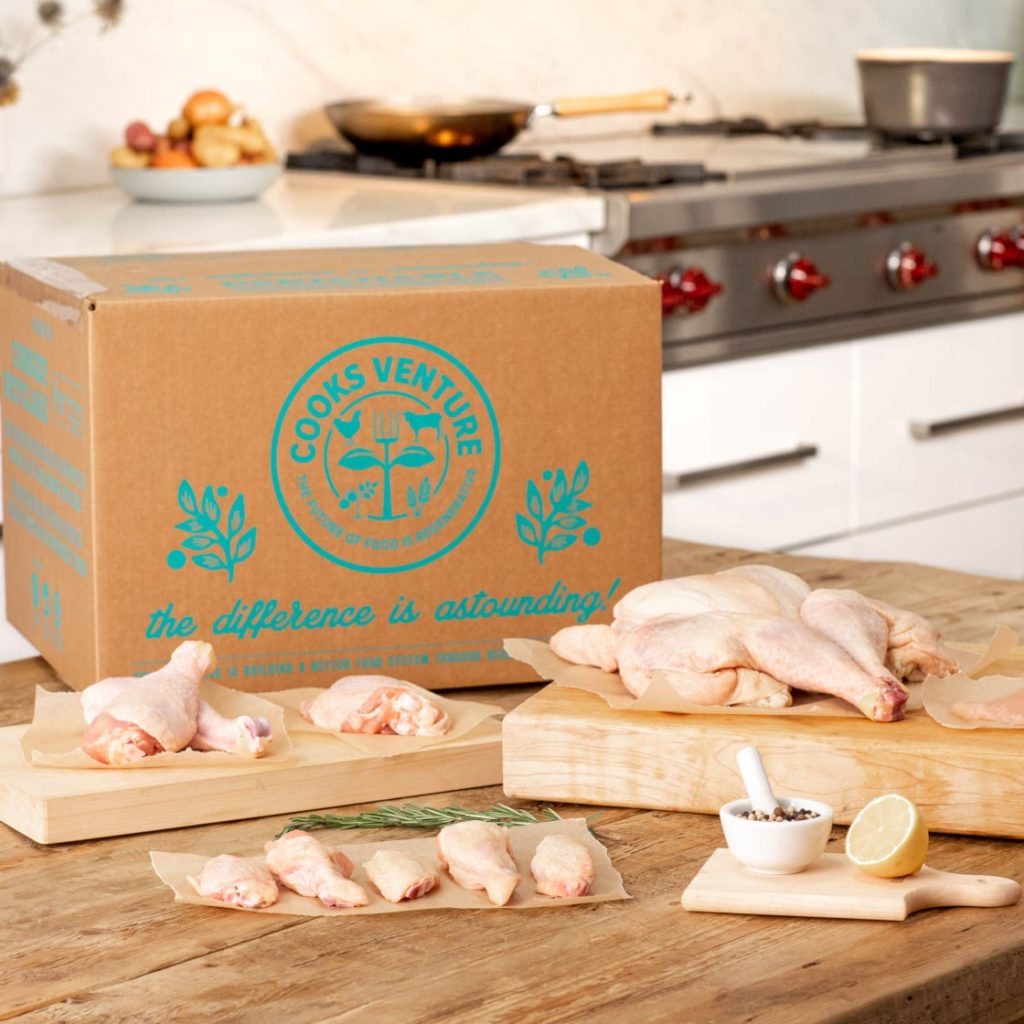 Raw chicken parts including drumsticks, wings, and a whole chicken displayed on parchment paper and wooden cutting boards on a kitchen counter next to a large cardboard box labeled 'Cooks Venture' in turquoise text, with a bowl of mixed peppercorns and a halved lemon nearby.