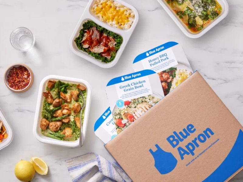 Blue Apron meal kit box partially open on a marble countertop with containers of prepared meals including grilled chicken, macaroni and cheese, greens with BBQ pulled pork, and a vegetable soup, alongside a glass of water, lemon halves, and a small bowl of red pepper flakes.