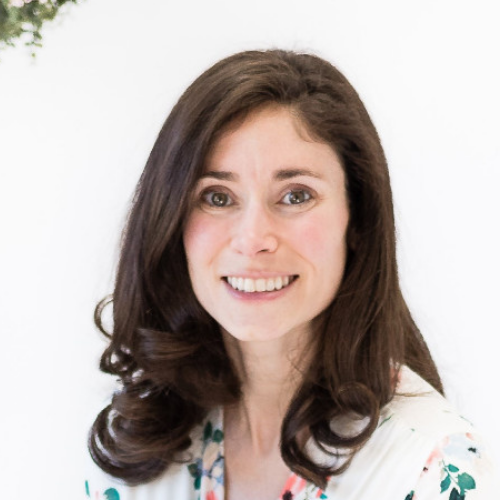 Smiling woman with dark brown hair styled in loose curls, wearing a white top with colorful floral patterns, against a plain white background.