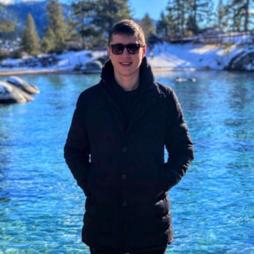 A man wearing dark sunglasses and a black winter coat stands with his hands in his pockets in front of a clear blue lake with snow-covered rocks and pine trees in the background.