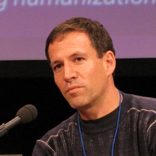 Man with short dark hair speaking into a microphone, wearing a dark sweater and a blue lanyard, against a blurred purple background.