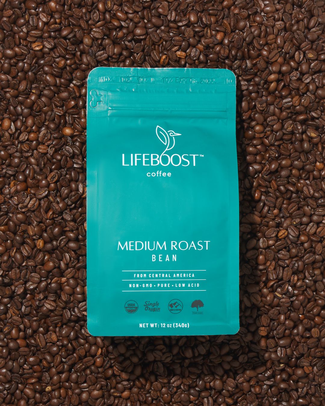 A teal-colored package of Lifeboost coffee labeled Medium Roast Bean, placed on a bed of roasted coffee beans. The package highlights attributes: from Central America, non-GMO, pure, low acid, USDA organic, single origin, high elevation, shade grown, and net weight 12 oz (340g).