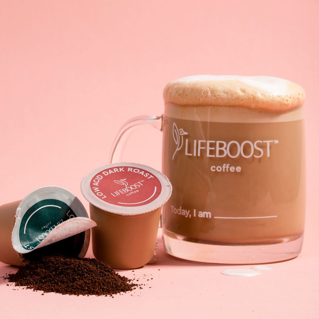 Clear glass mug of Lifeboost coffee with foam atop and text 'Today, I am ___' on a pink background next to two coffee pods, one open with coffee grounds spilled.