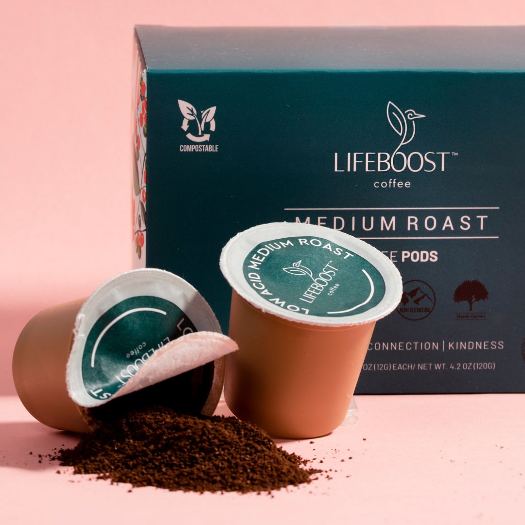 Two Lifeboost coffee medium roast single-serve pods on a pink surface, one tipped over with coffee grounds spilling out, and a Lifeboost coffee box in the background.