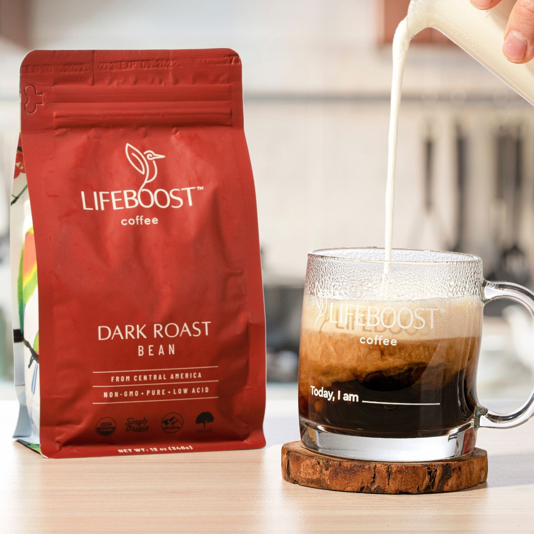 Bag of Lifeboost Dark Roast coffee beans next to a clear glass mug with Lifeboost coffee and text "Today, I am" filled with dark coffee as milk is poured into it, on a wooden surface.