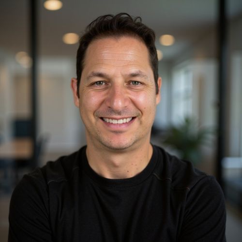 Smiling middle-aged man with short dark hair wearing a black shirt, standing indoors with blurred background of a modern office space.