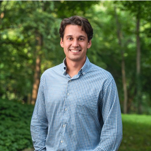 Smiling young man wearing a blue checkered shirt standing outside with trees and greenery in the background.