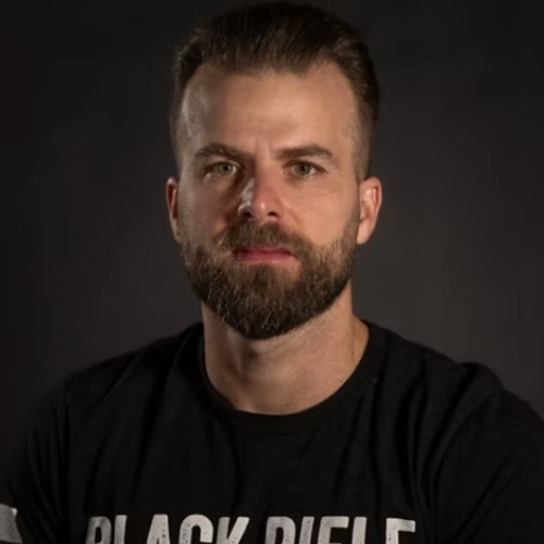 Portrait of a man with a beard and mustache wearing a black t-shirt with partial visible text reading 'BLACK RIFLE' against a dark background.