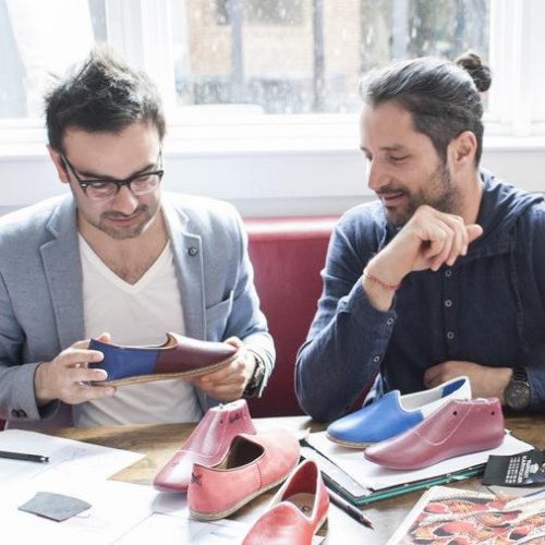 Two men sitting at a table examining colorful shoe prototypes and shoe samples in a bright room with large windows.