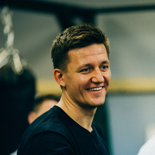 Smiling man with short brown hair wearing a dark shirt, standing indoors with blurred background and people.