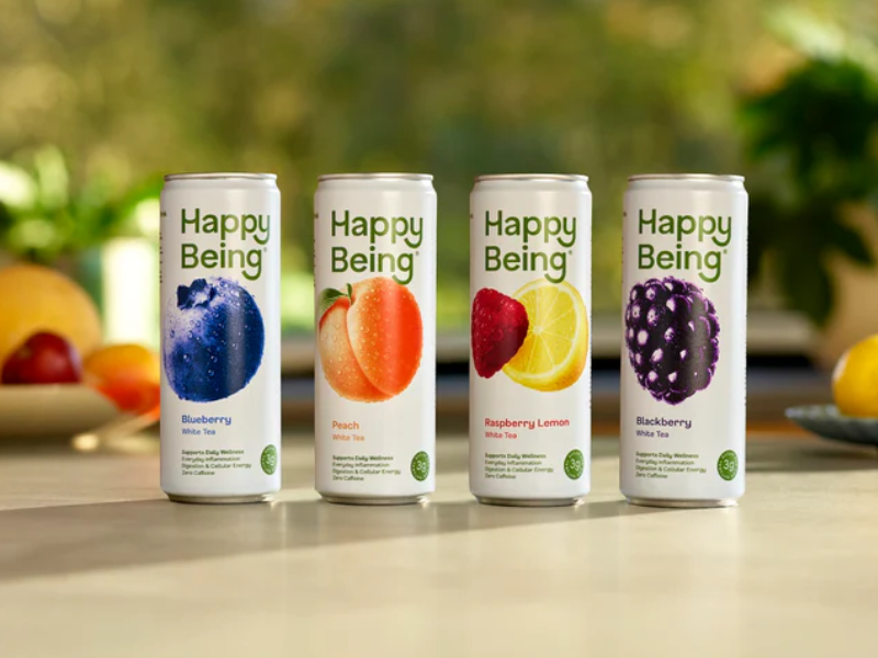 Four tall cans of Happy Being white tea lined up side by side on a light-colored table with blurred greenery in the background, flavors include Blueberry, Peach, Raspberry Lemon, and Blackberry, each featuring large fruit images on the cans.