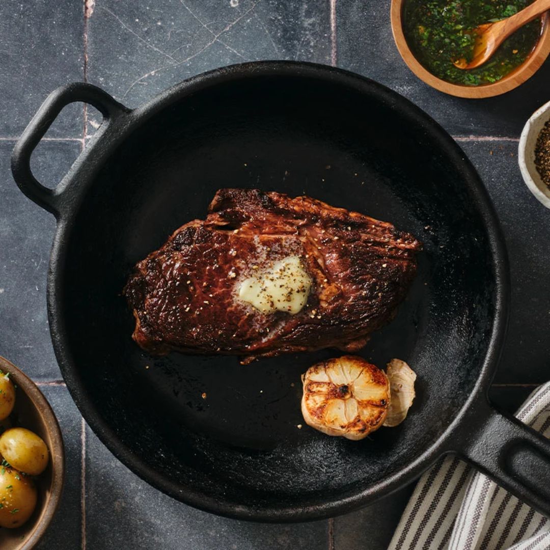 Skillet with a cooked steak topped with melting butter and seasoned with pepper, alongside a roasted garlic head, on a dark tiled surface with side bowls of small potatoes and green sauce visible.