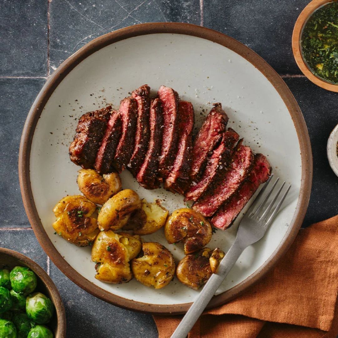Plate with sliced medium-rare steak and roasted golden-brown gnocchi, a fork on the side, with side dishes of Brussels sprouts and green sauce visible around the plate on a dark tiled surface and an orange cloth napkin.