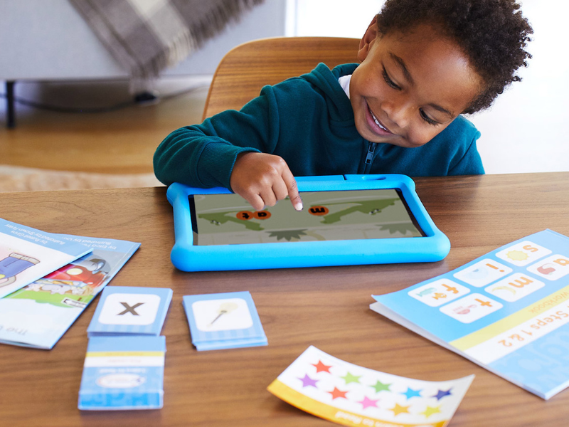 Child smiling and pointing at a blue tablet showing an educational game with letters on the screen, surrounded by flashcards and learning materials on a wooden table.