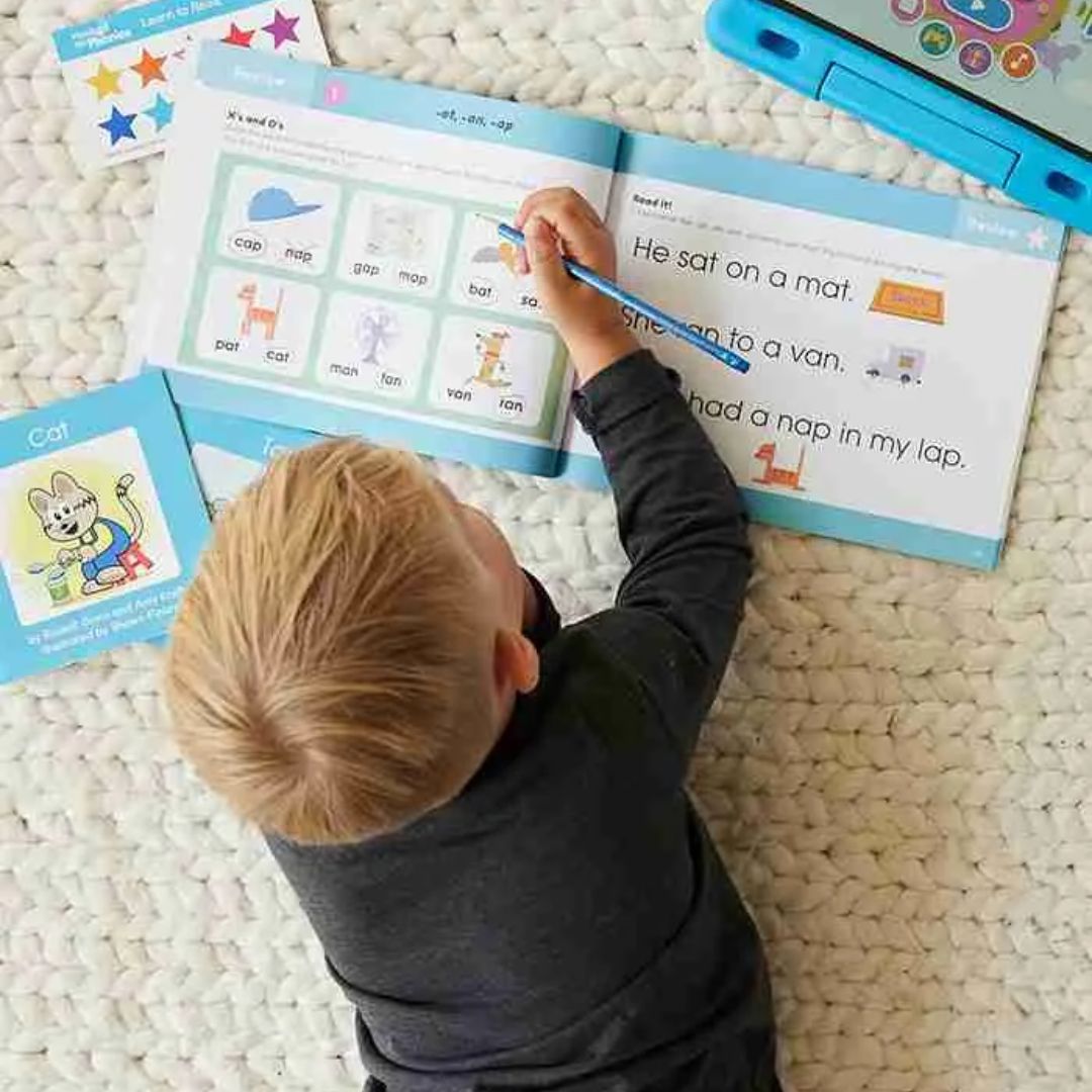 Child lying on a textured cream rug writing in a children's workbook with a blue colored pencil, surrounded by flashcards and a blue tablet.