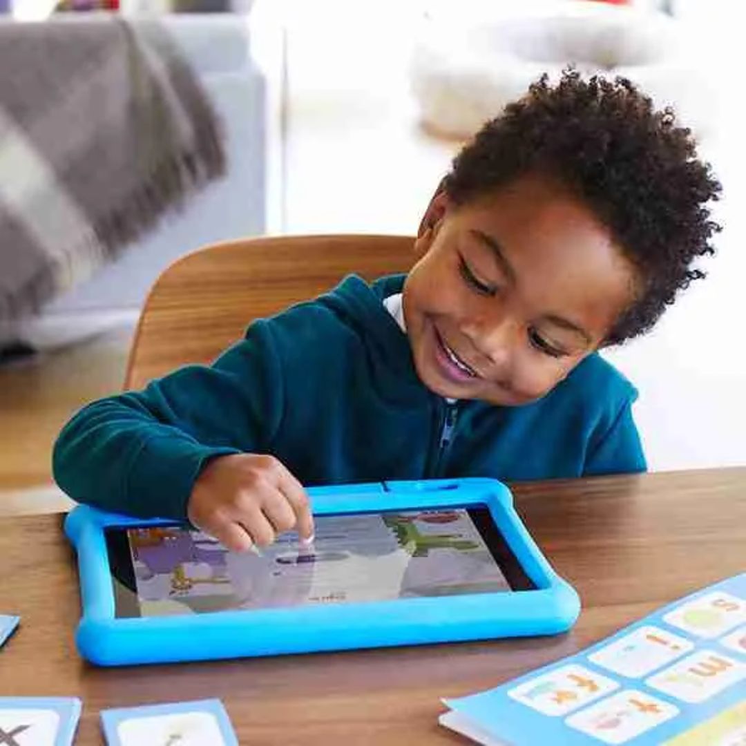 Young child with curly hair smiling and interacting with a blue-cased tablet on a wooden table, surrounded by educational flashcards.
