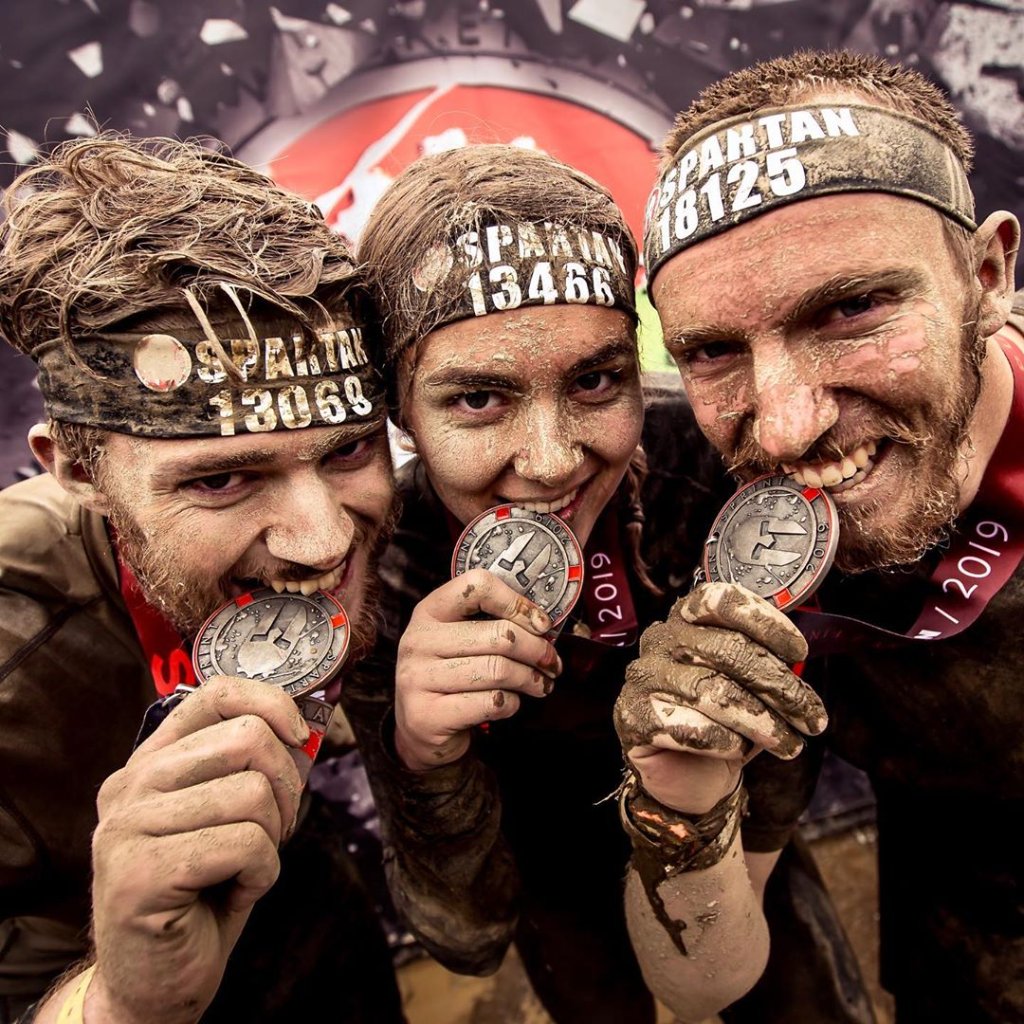 Three muddy people wearing Spartan headbands with numbers, smiling and biting their Spartan race finisher medals with red ribbons.