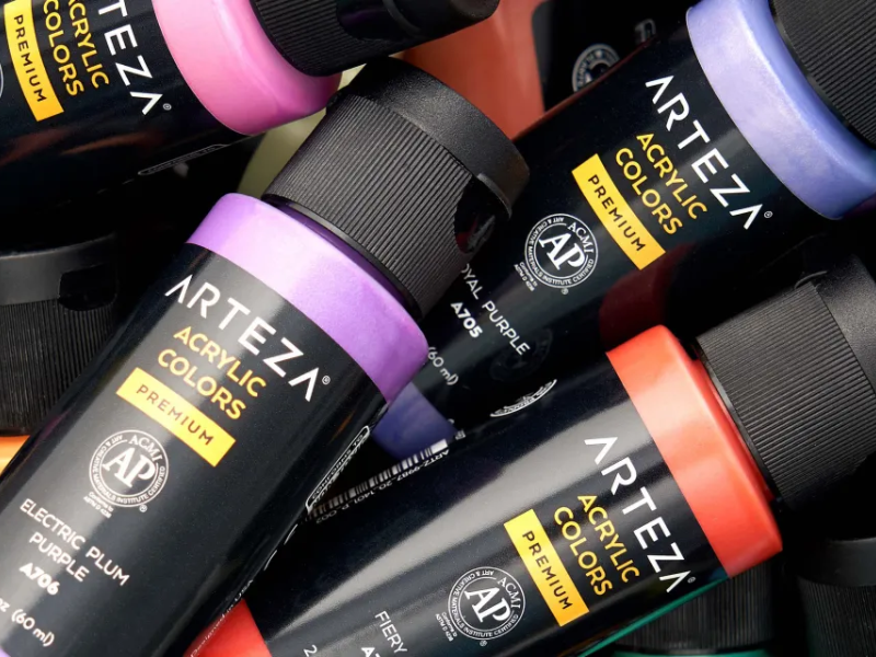 Close-up of five Arteza brand acrylic paint tubes in various colors including electric plum purple, royal purple, and fiery red, with black caps and black labels featuring yellow and white text.