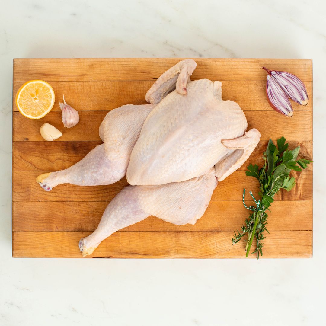 Whole raw chicken spatchcocked and laid flat on a wooden cutting board, surrounded by half a lemon, two garlic cloves, two halved shallots, and a small bunch of fresh herbs including rosemary and parsley.