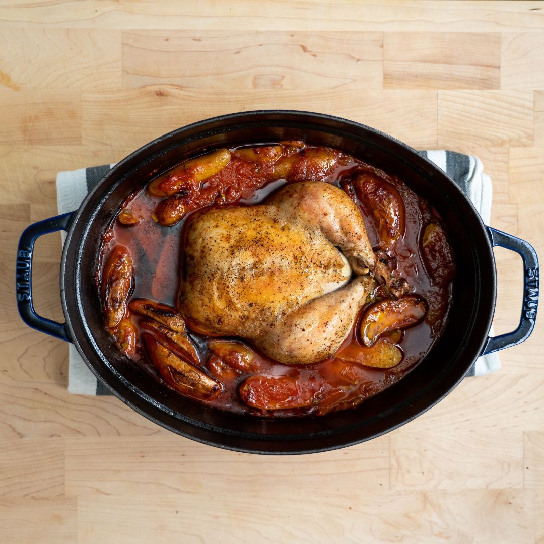 Whole roasted chicken seasoned with pepper in a black oval Staub cookware on a wooden surface, surrounded by roasted vegetables in a red sauce.