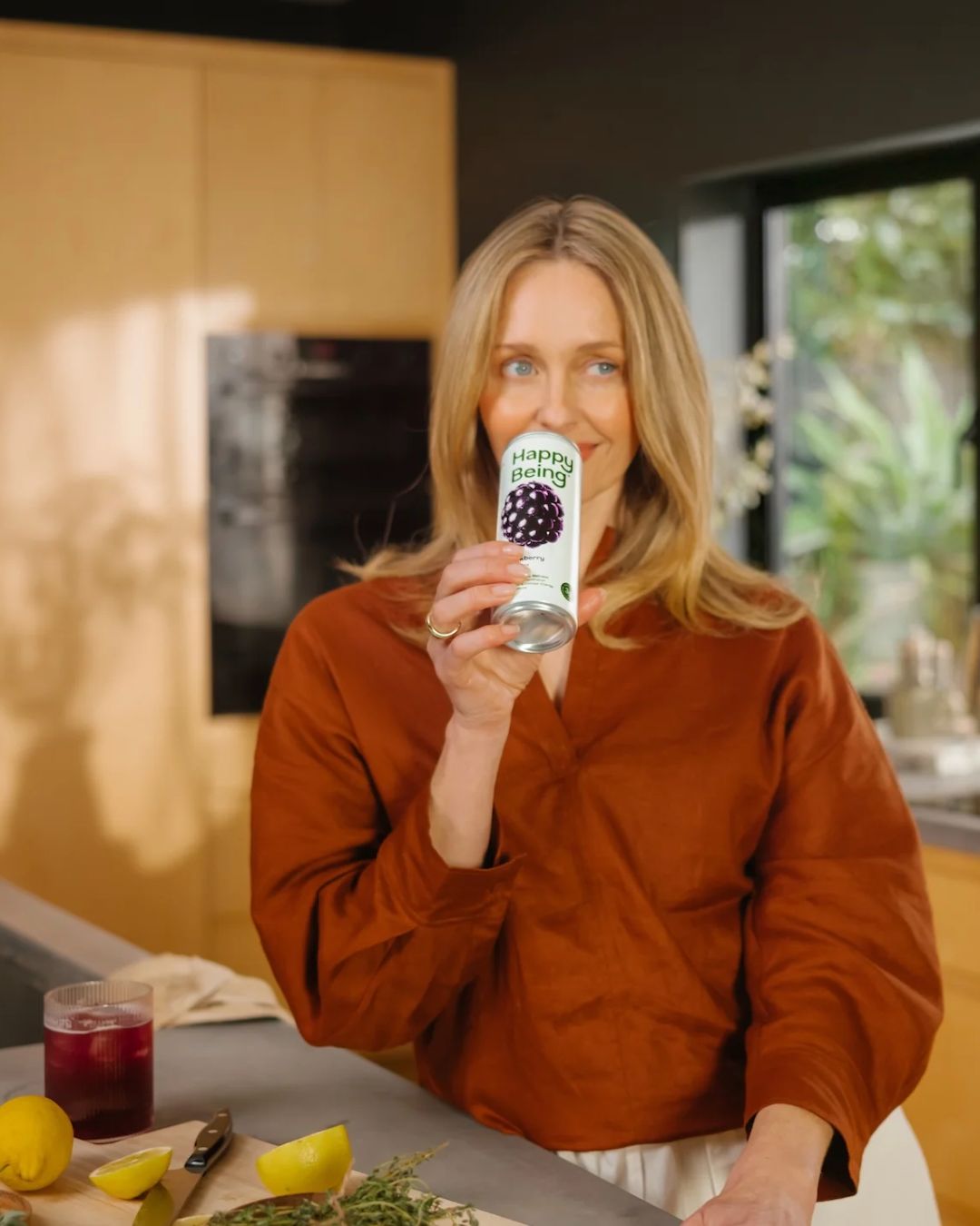 Smiling woman with blonde hair wearing a rust-colored blouse standing in a kitchen holding a can of Happy Being blackberry drink near her mouth. On the counter are lemons, a knife, herbs, a glass with a dark red beverage, and a cloth.