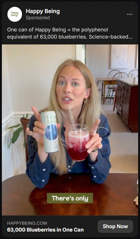 A woman with long blonde hair sitting at a table holding a can of Happy Being and a glass with a red drink and ice, with a straw, promoting the health benefits of the drink equivalent to 63,000 blueberries.