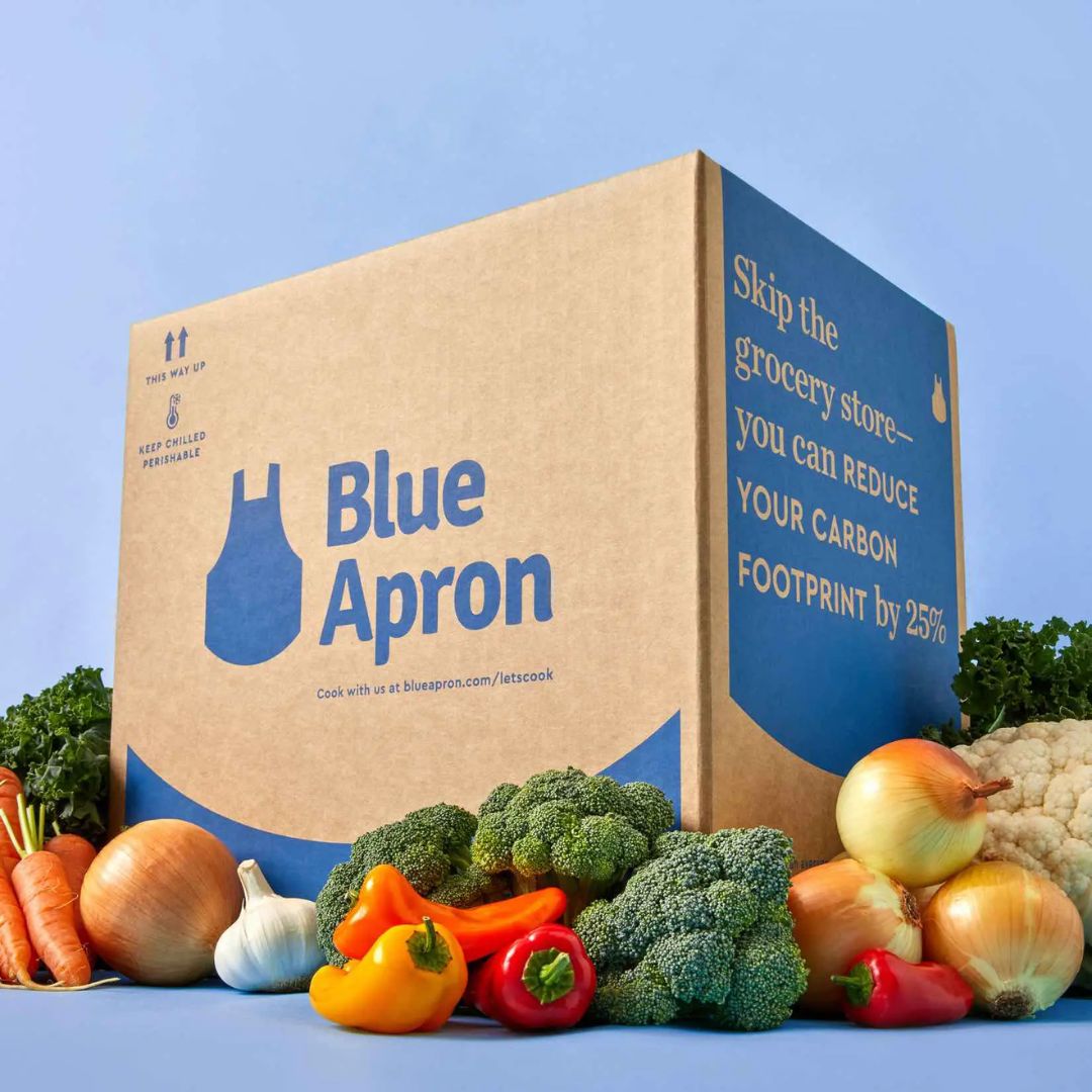 A Blue Apron delivery box surrounded by fresh vegetables including carrots, onions, garlic, broccoli, bell peppers, cauliflower, and kale, against a blue background.