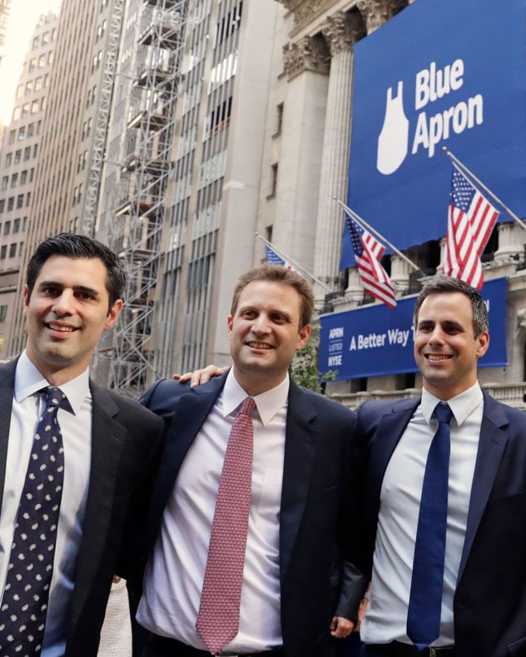Three men in business suits smiling and posing together in front of a building with American flags and a large blue sign reading 'Blue Apron' and 'A Better Way'.