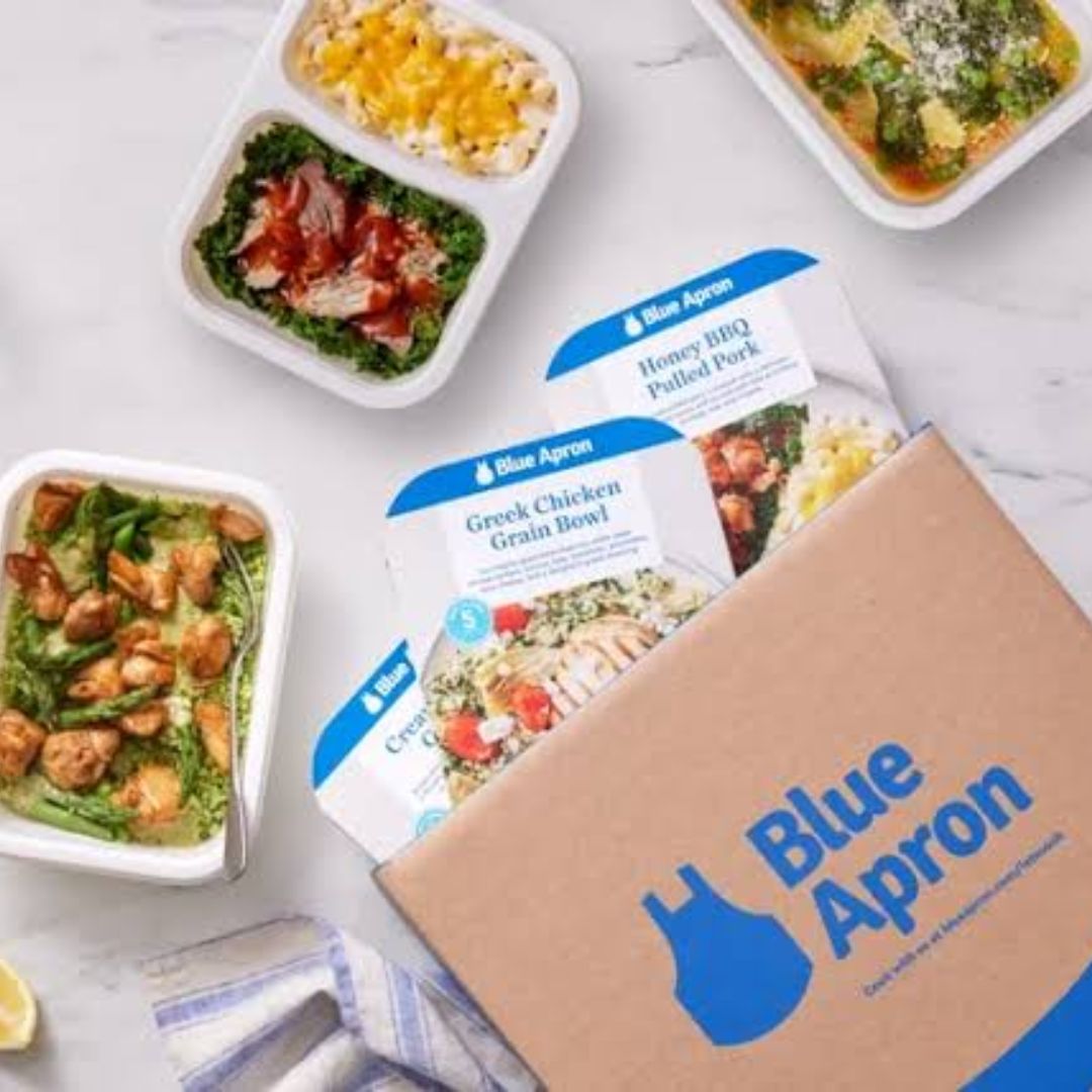 Blue Apron meal kit box partially open showing recipe cards for Honey BBQ Pulled Pork and Greek Chicken Grain Bowl surrounded by prepared meal containers with chicken, greens, macaroni and cheese, and other dishes on a marble surface.