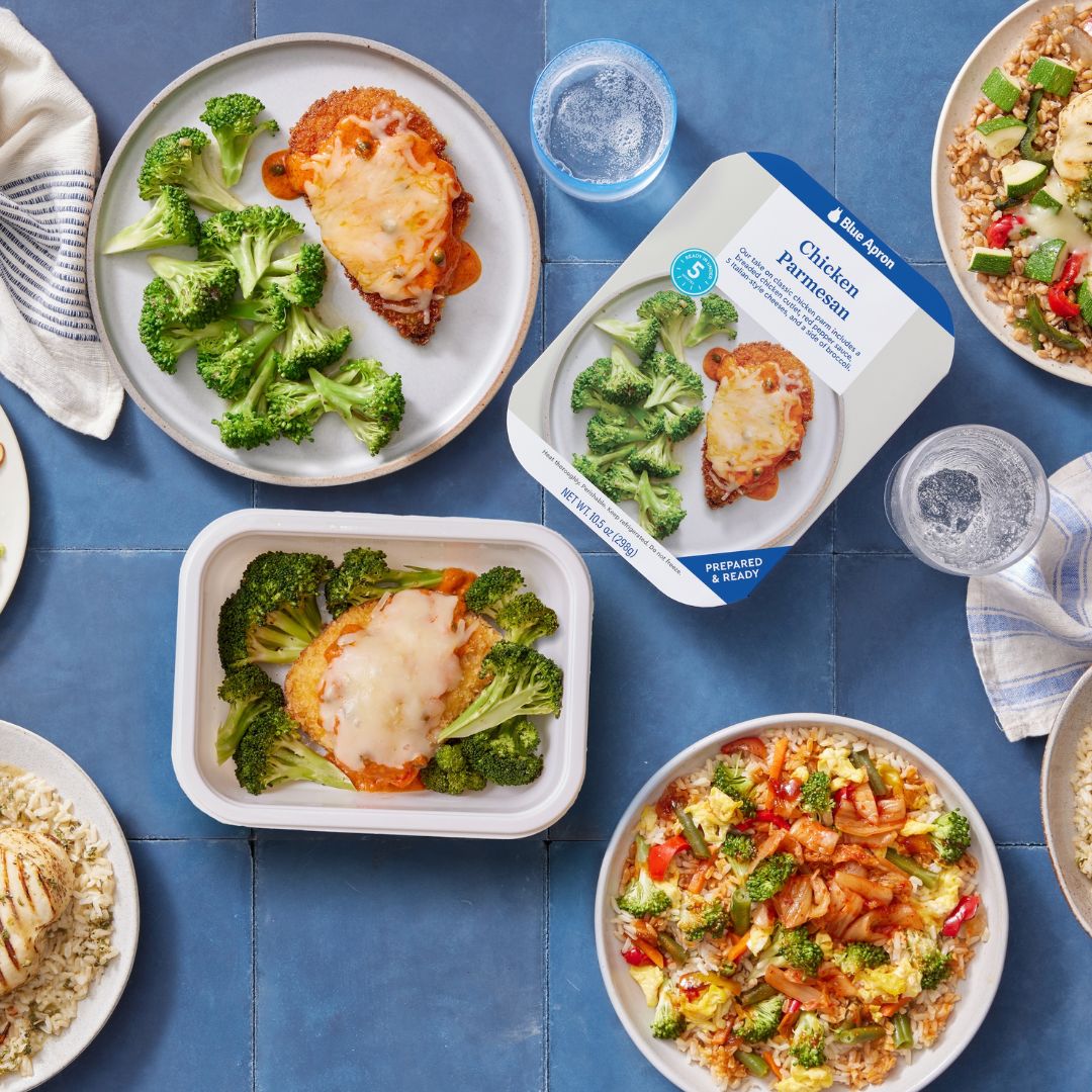 A Blue Apron meal kit featuring chicken parmesan topped with melted cheese and a side of steamed broccoli, displayed on a plate and in its packaging, surrounded by other prepared dishes on a blue tiled surface.