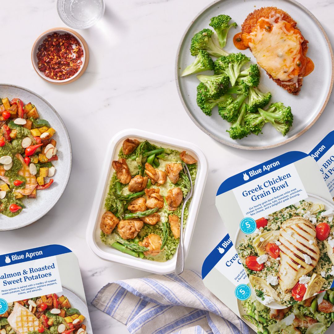 Overhead view of three plated meals and food containers on a white marble surface with a striped cloth napkin. A plate holds broccoli florets and a breaded chicken cutlet topped with melted cheese and sauce; another plate has salmon with roasted sweet potatoes, bell peppers, and pesto sauce garnished with almond slices; a white container contains pieces of grilled chicken with asparagus on a green sauce and grain base. Blue Apron labels for Greek Chicken Grain Bowl and Salmon & Roasted Sweet Potatoes visible.