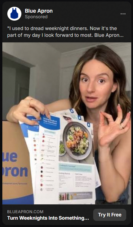 A woman holding and showing Blue Apron meal recipe cards with a prepared dish photo, gesturing with her hand as if explaining or emphasizing something.