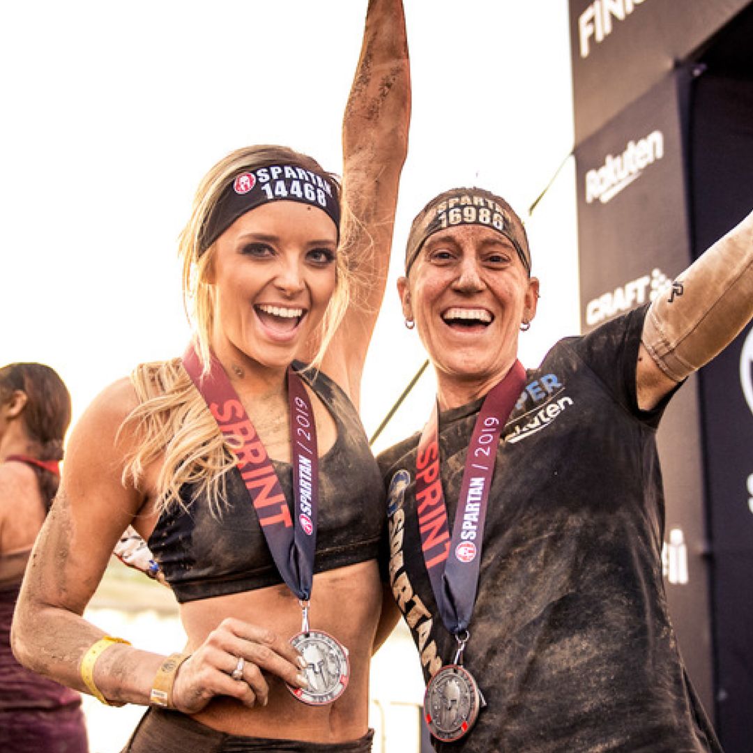 Two smiling women covered in mud wearing Spartan headbands and medals around their necks, celebrating their finish in a race.