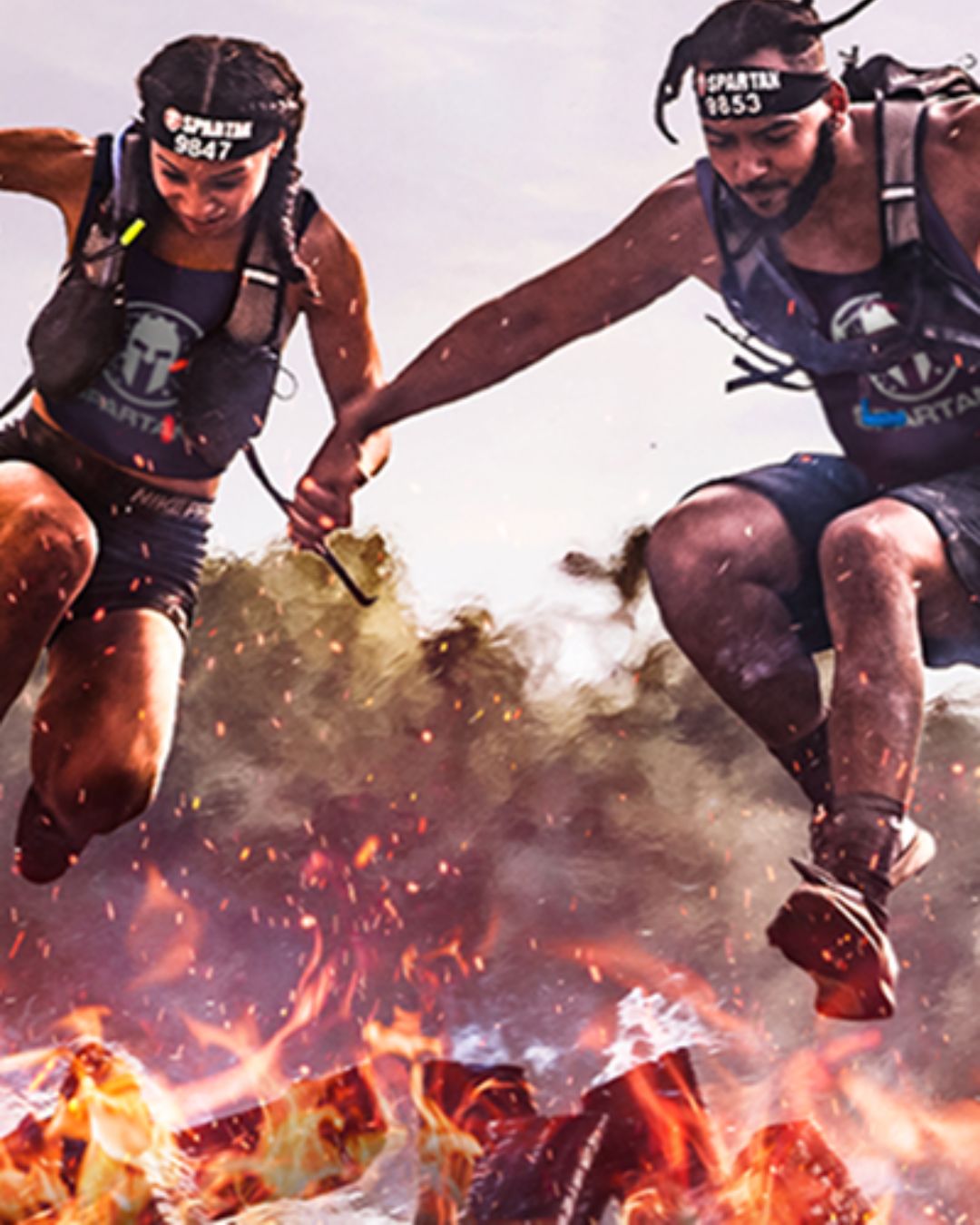 Two athletes wearing Spartan headbands and gear holding hands as they jump over flames and smoke in an intense obstacle race.