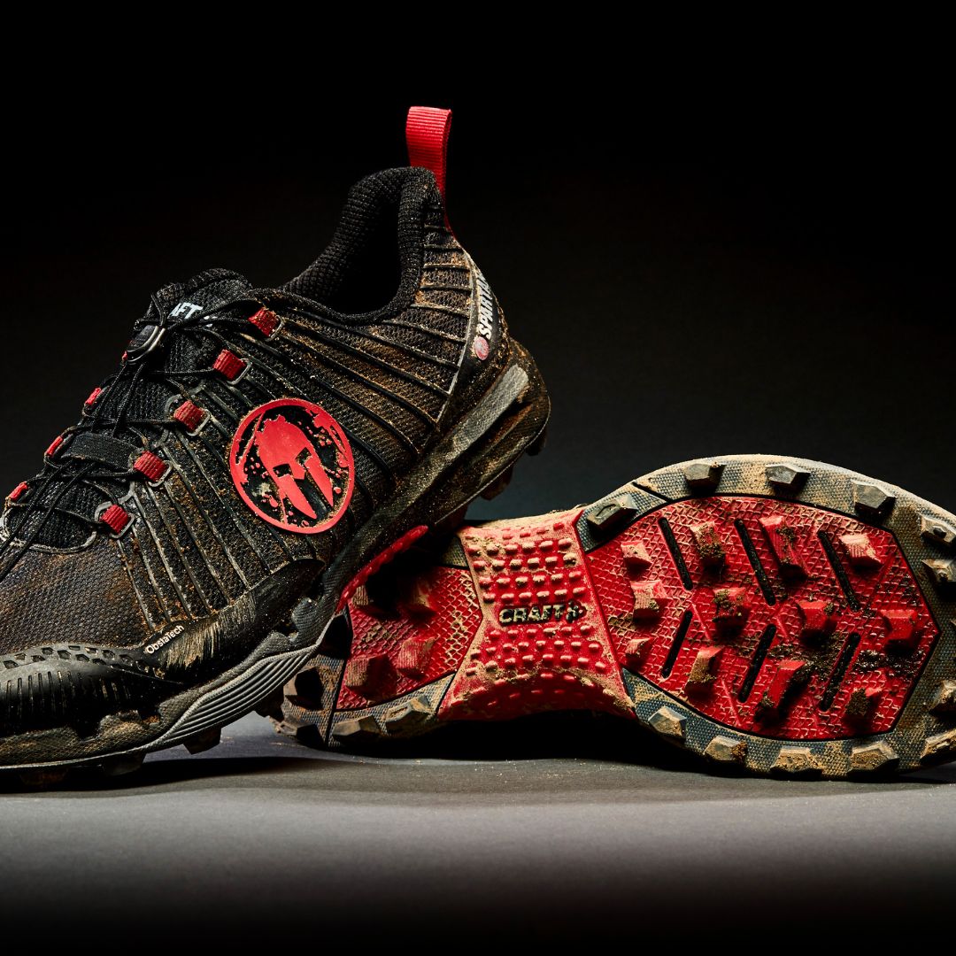 Pair of black and red Spartan race shoes with rugged tread and dirt on soles, displayed against a black background.
