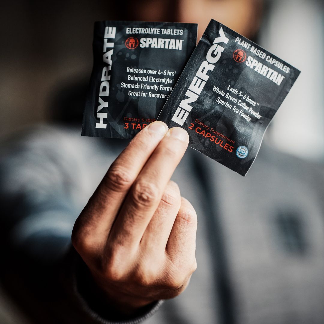 Hand holding two Spartan branded supplement packets: one labeled Hydrate electrolyte tablets with 3 tablets, and the other labeled Energy plant-based capsules with 2 capsules.
