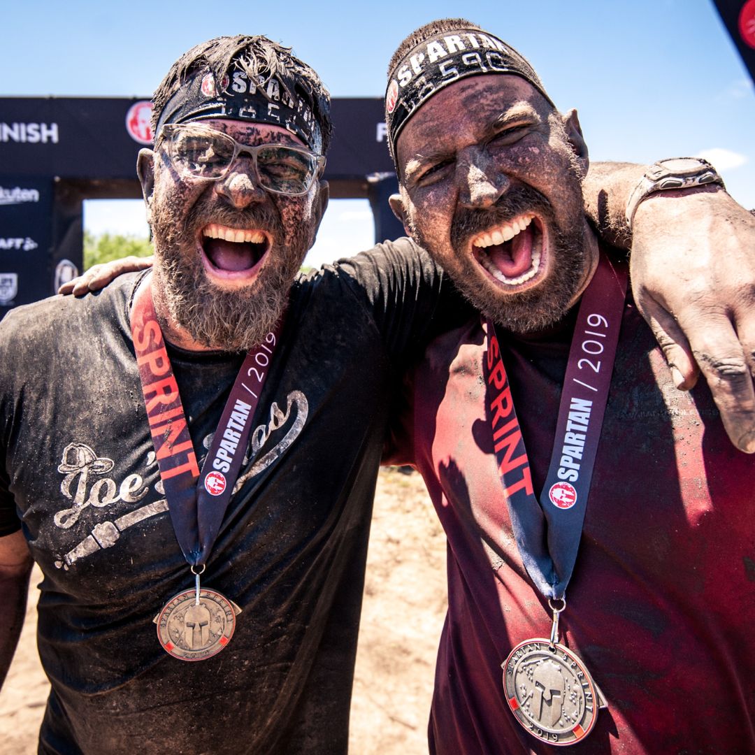 Two men covered in mud with Spartan headbands and Sprint 2019 finisher medals around their necks, celebrating with arms around each other and wide joyful smiles.