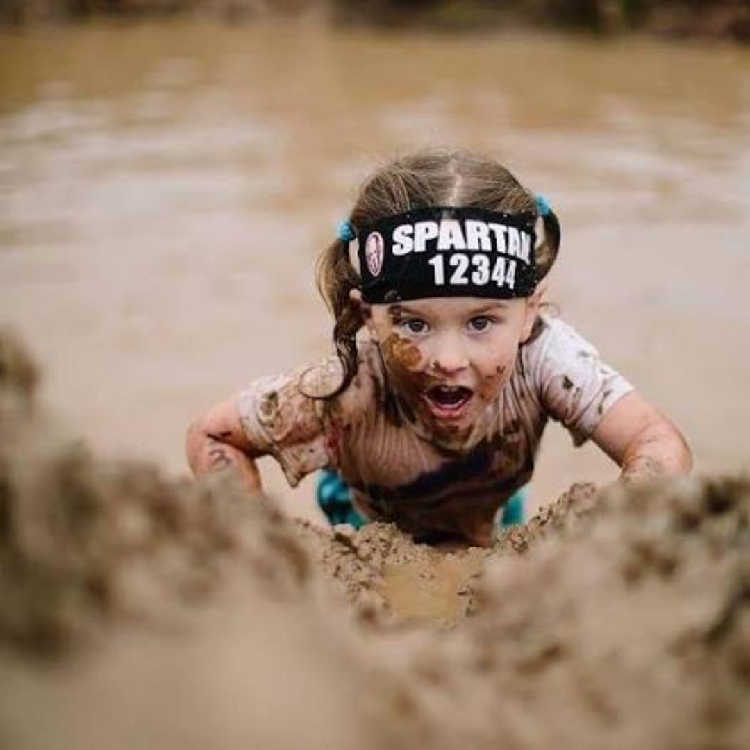A young girl wearing a black headband labeled 'SPARTAN 12344' climbs through muddy terrain with mud on her face and clothes, showing determination.