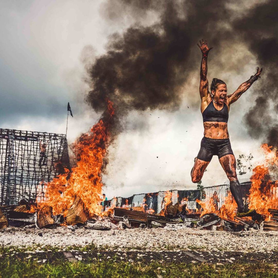 Athlete covered in mud wearing black sports bra and shorts, jumping excitedly over a fire pit with large flames and thick black smoke during an outdoor obstacle race.
