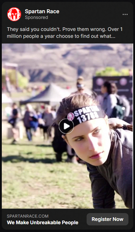 Woman participant in a Spartan Race wearing a headband with the number 13708 crouching on grass with a mountainous background and blurred people behind her.