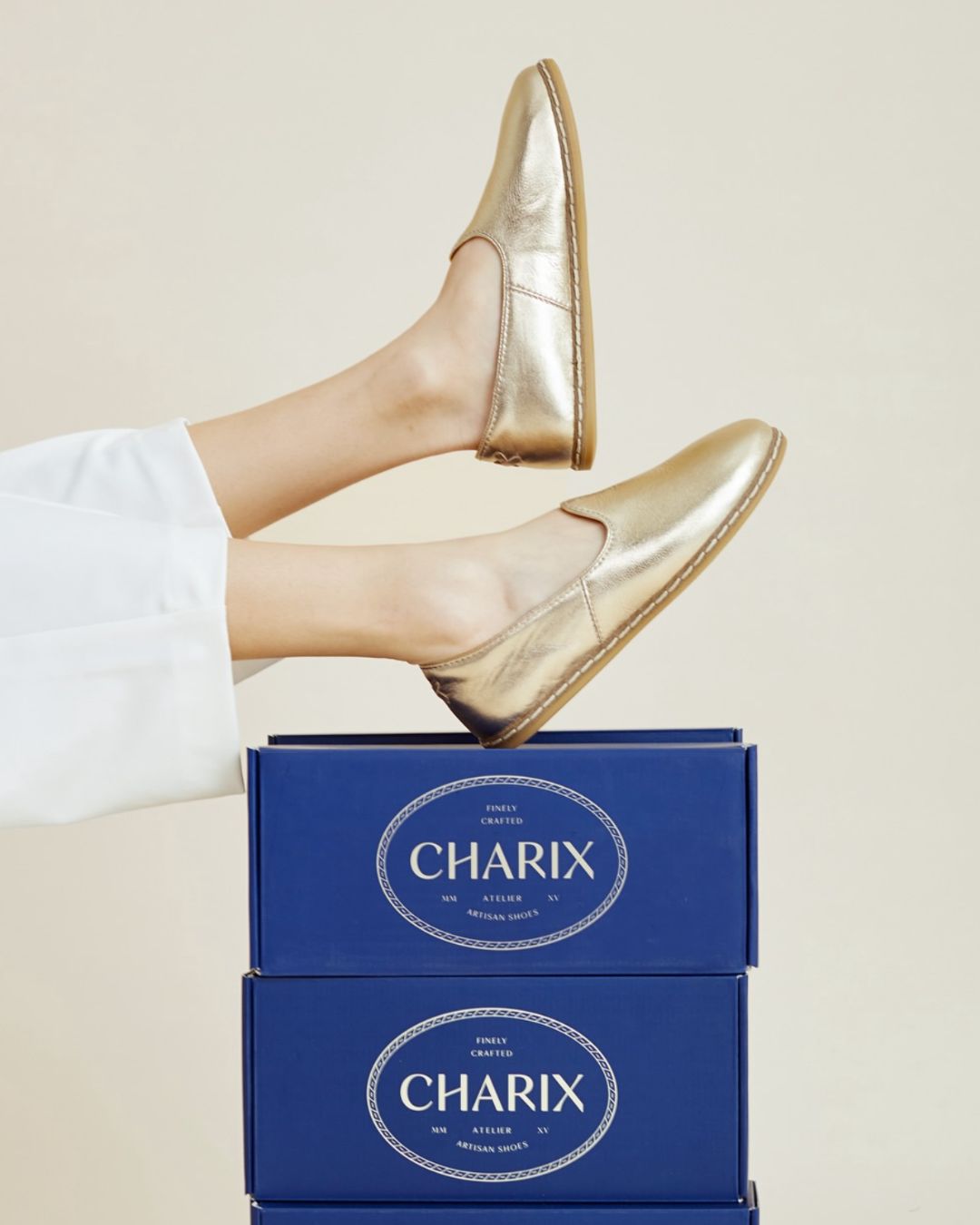 Pair of gold slip-on shoes worn on feet raised above stacked blue Charix branded shoeboxes against a beige background.