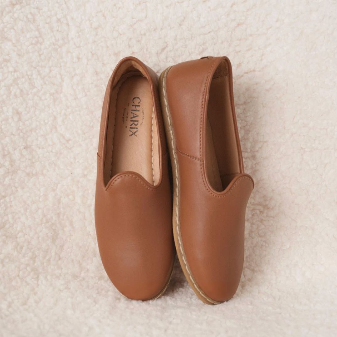 Pair of brown slip-on leather shoes with stitching details, placed side by side on a soft, light textured surface.