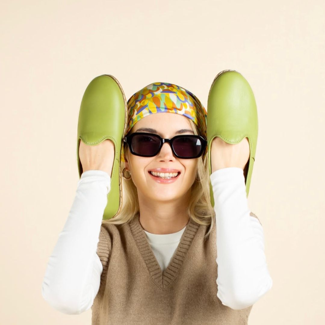 Smiling woman wearing dark sunglasses and a colorful headscarf holds up a pair of green slip-on shoes near her face, wearing a beige sleeveless sweater over a white long-sleeve shirt against a light beige background.