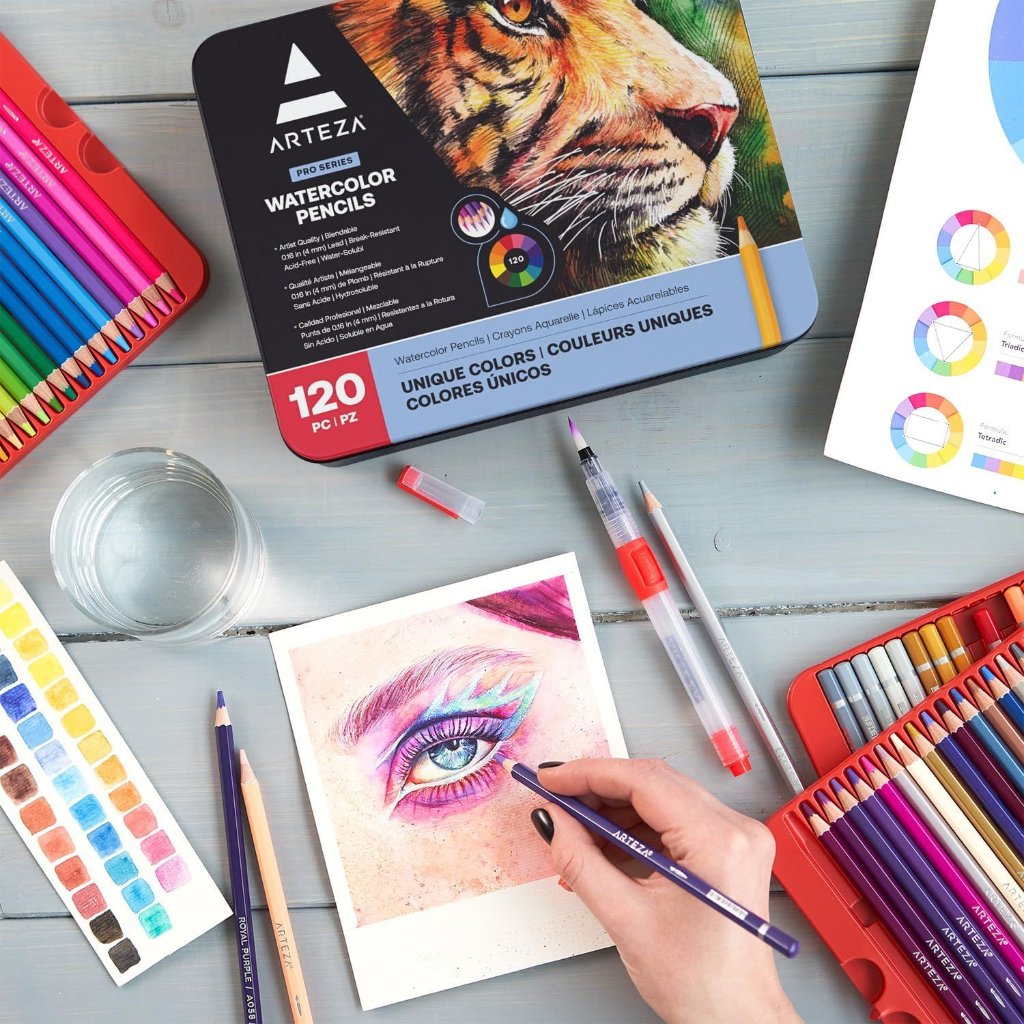 A hand with black nail polish coloring a detailed eye drawing on paper using a purple Arteza watercolor pencil, surrounded by an open set of colored Arteza watercolor pencils, a water brush, a glass of water, color swatches, and a color theory chart on a wooden surface.