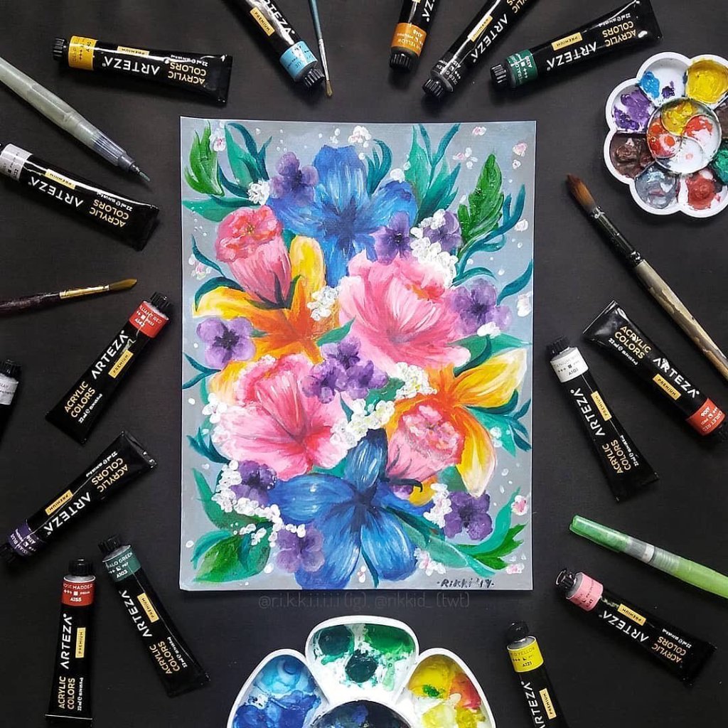 Colorful acrylic painting of a floral bouquet with pink, blue, orange, and purple flowers surrounded by green leaves on a gray background, with paint tubes, brushes, and palettes around it.