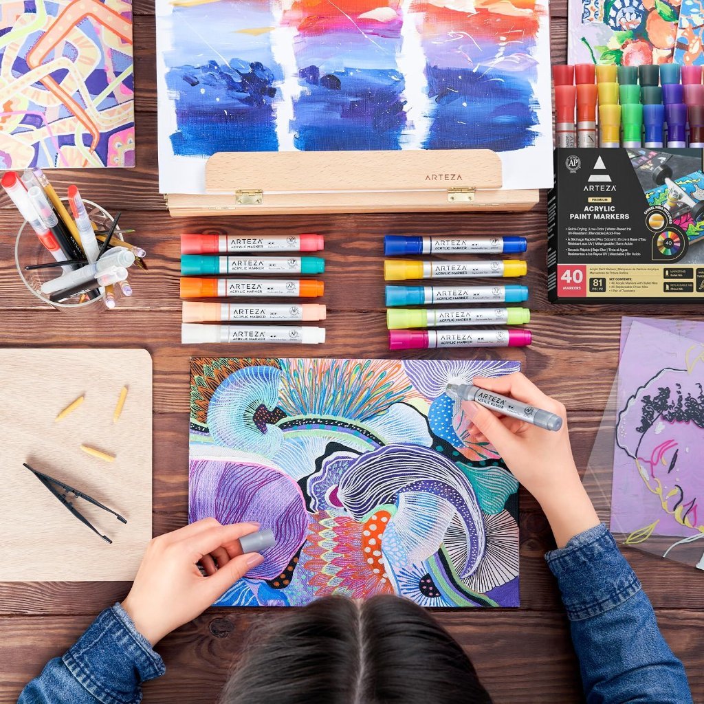 Person coloring an intricate abstract design with Arteza acrylic paint markers on a wooden table surrounded by colorful markers, paint samples on a wooden easel, and a small cup of brushes and tools.