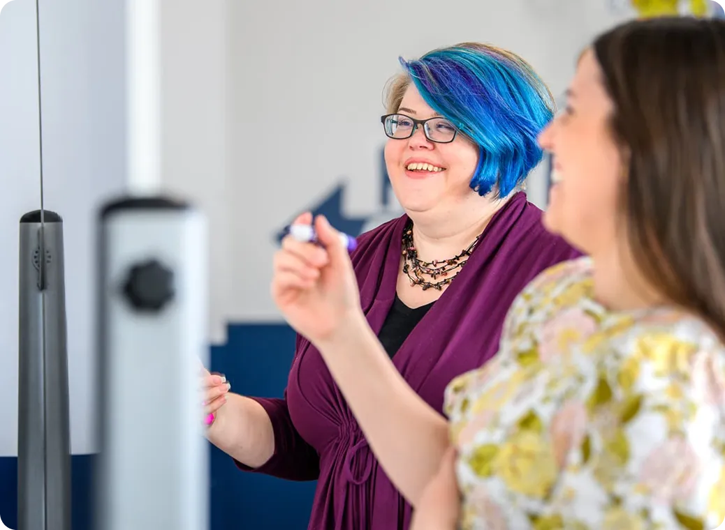 Two women smiling and interacting in a casual setting; one has blue and purple hair, glasses, a purple top, and a necklace, while the other woman, out of focus in the foreground, wears a floral shirt.