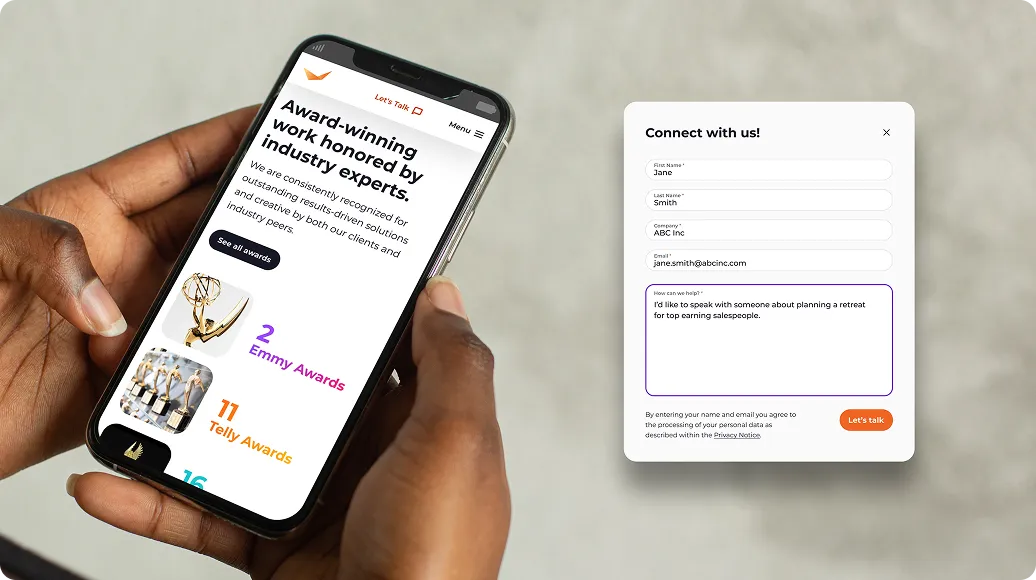 Hand holding a smartphone displaying a website announcing award-winning work recognized by industry experts with Emmy and Telly awards, alongside a contact form on the right side titled 'Connect with us!' with fields filled with sample information for name, company, email, and a message about planning a retreat for top salespeople.