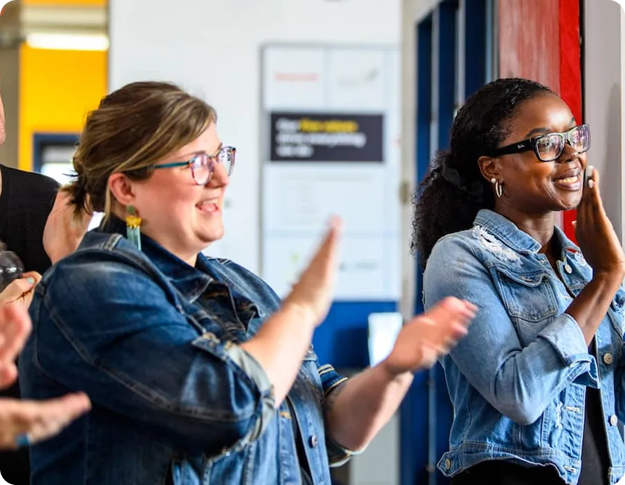 Two women smiling and clapping indoors, both wearing denim jackets and glasses, with a blurred background of a sign and door.