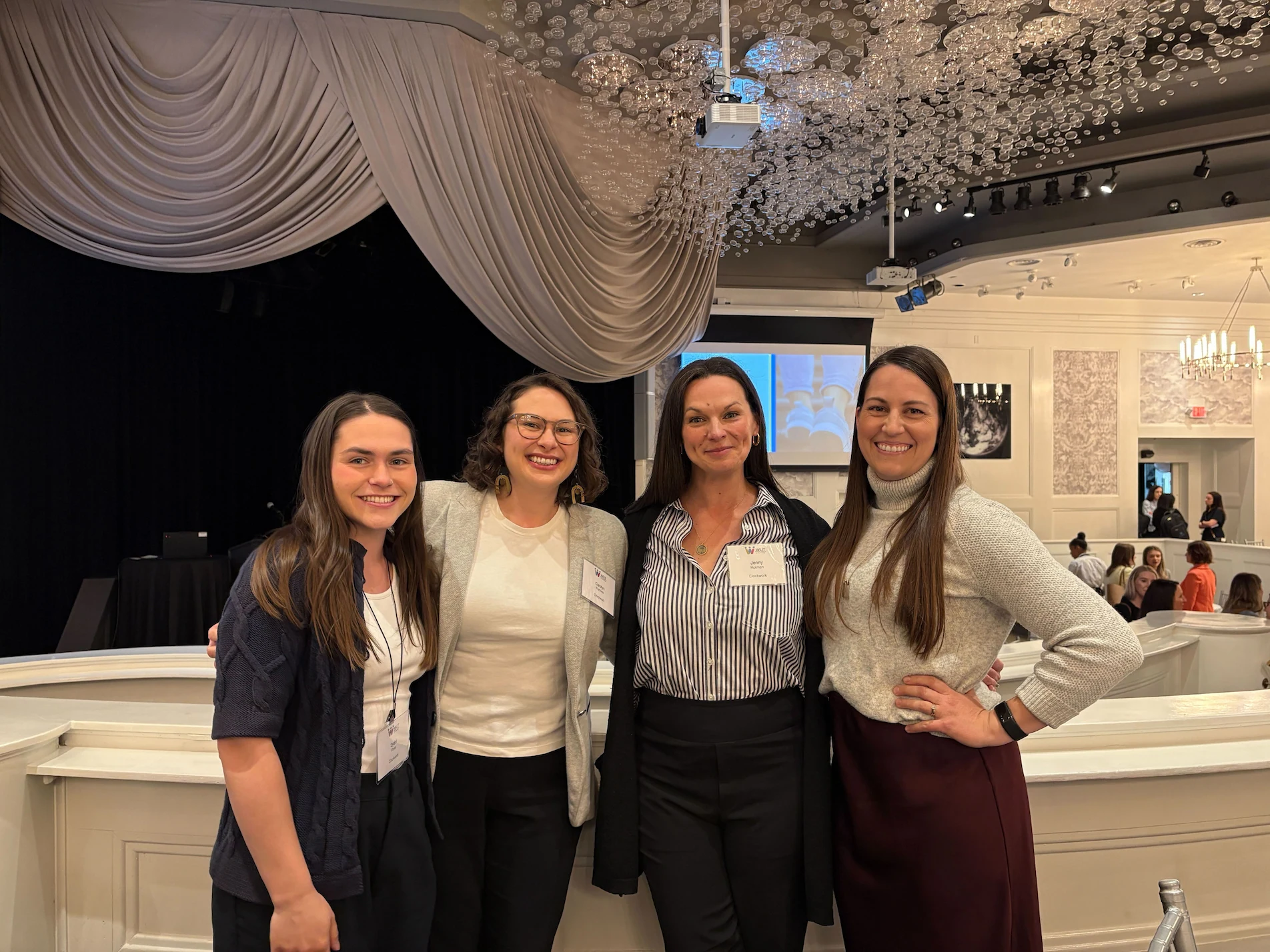 Four women standing close together inside a formal event space with draped ceiling and elegant lighting, smiling toward the camera, two wearing name tags.